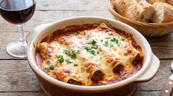 Fuente de cerámica con canelones horneados cubiertos de queso y salsa de tomate, junto a una copa de vino tinto y una cesta con pan.