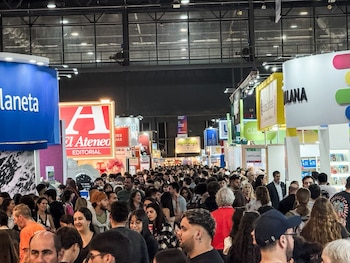 Multitud de personas en un pasillo de una convención interior. Grandes banners de "Planeta", "El Ateneo Editorial" y "Santillana" cuelgan sobre los stands