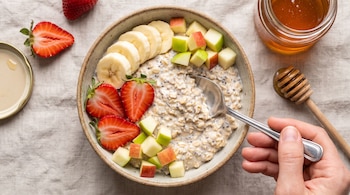 Bol con avena, chía, rodajas de banano, fresas cortadas y cubos de manzana. Una mano acerca una cuchara a la mezcla. Un tarro de miel y un cucharón de madera.