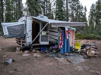 Un camper blanco con toldo extendido, una extensión de lona y ropa tendida, estacionado en un terreno de tierra con rocas y árboles