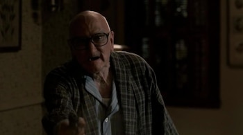 Primer plano de un hombre calvo y mayor con gafas y bata de cuadros, que gesticula con la mano derecha. Tiene la boca abierta y parece estar hablando