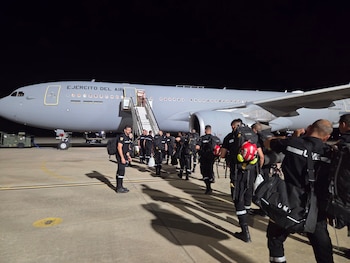 Avión gris del Ejército del Aire estacionado de noche en una pista, con escaleras. Personal de la UME con uniformes y mochilas embarca, uno con casco rojo