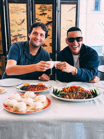 Dos hombres sentados a una mesa con platos de pescado, bollos al vapor y tazas pequeñas, frente a un biombo con ilustraciones de peces