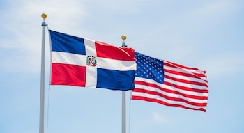 Dos banderas, una de República Dominicana con su escudo y otra de Estados Unidos con estrellas y barras, ondean sobre astas bajo el cielo azul.