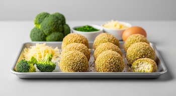 Una bandeja metálica con croquetas de brócoli, algunas piezas de brócoli fresco, queso rallado, un recipiente con hierbas picadas y un huevo.