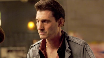 Primer plano del actor Robert Funaro, con cabello oscuro peinado hacia atrás, luciendo una camisa burdeos abierta y una chaqueta gris