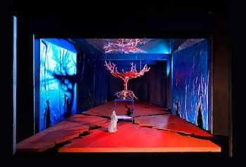 Maqueta de escenografía con un árbol rojo central, un lecho azul, una figura blanca y un escenario fracturado, iluminado en tonos azul y rojo