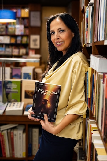 Una mujer sonriente de cabello oscuro sostiene un libro junto a estanterías con libros en una librería