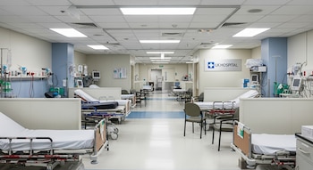 Interior de un pasillo de hospital con camillas, sillas, monitores y postes de suero a ambos lados. Un letrero en la pared indica LK Hospital.