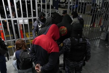 Agentes con chalecos tácticos y pasamontañas custodian a individuos con capuchas y esposas cerca de una reja metálica y vehículos