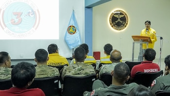 Un expositor habla desde un podio frente a un auditorio con bomberos, militares y personal con uniformes amarillos en una sala de conferencias