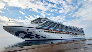 Un crucero blanco con el nombre Crown Princess y acentos azules en el agua. Se observa un muelle en primer plano y un cielo nublado