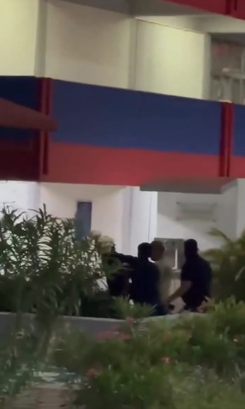 Un grupo de personas vestidas de oscuro rodea a un individuo frente a un edificio con paneles azules y rojos detrás de varias plantas