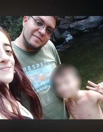 Una mujer y un hombre con gafas posan sonrientes junto a un niño que hace la señal de la paz, con rocas y un río detrás