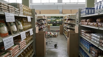 Pasillo de supermercado dominicano. Estantes con arroz, aceite, pan, leche, huevos, verduras y precios visibles. Una persona empuja un carrito de compras medio lleno.