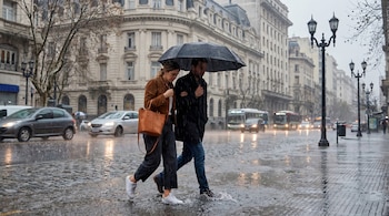 Un hombre y una mujer caminan bajo un paraguas negro en una calle adoquinada de Buenos Aires, en un día de lluvia intensa con charcos y edificios antiguos de fondo.