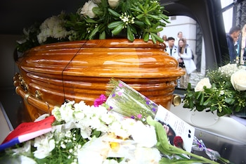 Un ataúd de madera barnizada con arreglos florales blancos y una tarjeta recordatoria en primer plano dentro de un vehículo