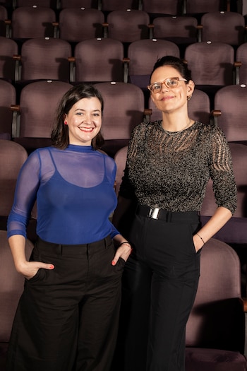 Primer plano de dos mujeres sonrientes de pie. La de la izquierda lleva una blusa azul, la de la derecha una blusa brillante. Detrás, asientos de teatro vacíos