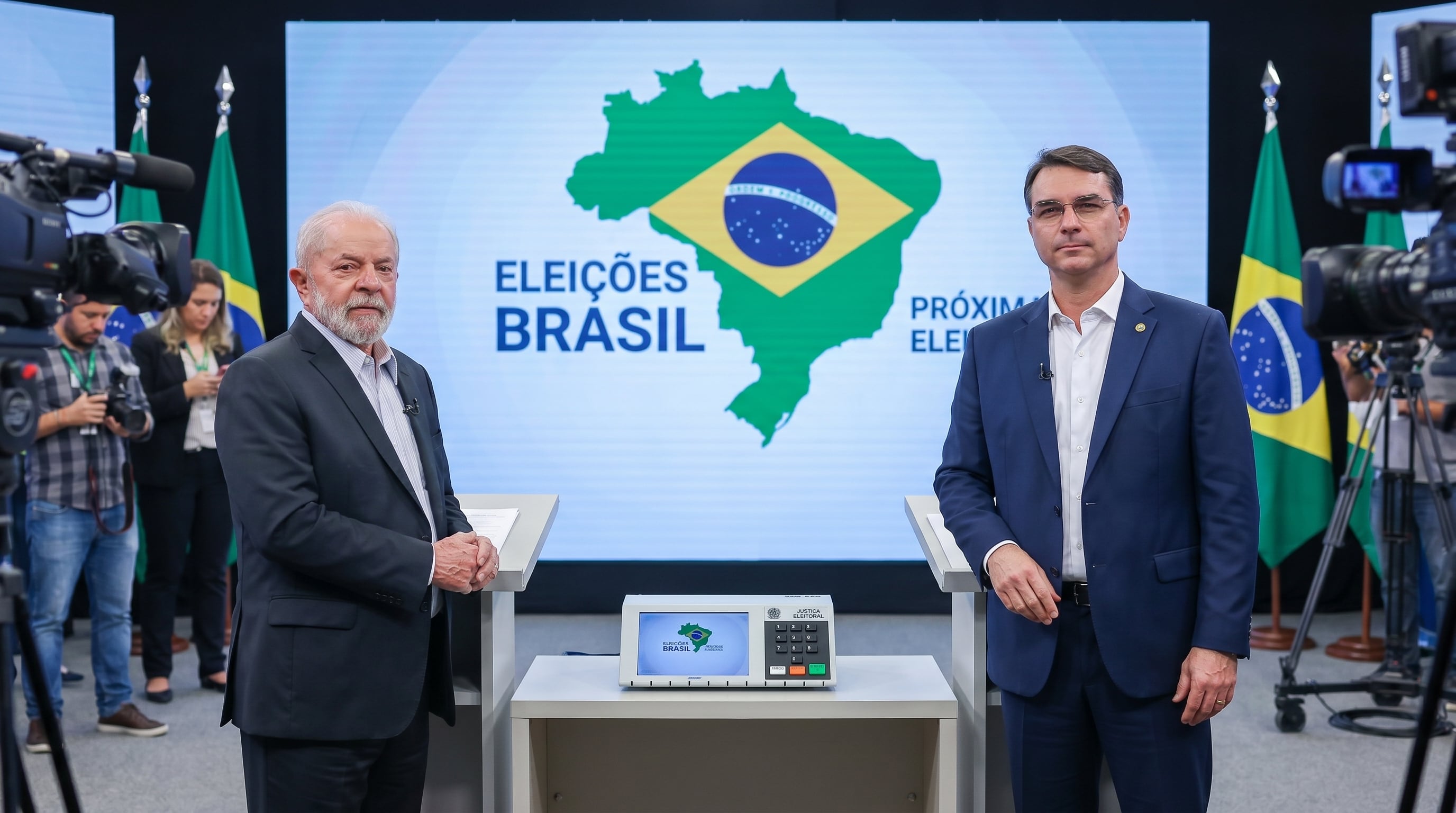 Lula da Silva y Flávio Bolsonaro posan en un evento electoral en Brasil, con una urna electrónica y el mapa del país de fondo, previo a las próximas elecciones. (Imagen Ilustrativa Infobae)
