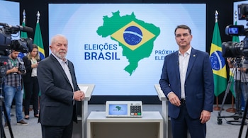 Lula da Silva y Flávio Bolsonaro de pie, vestidos con traje, frente a una pantalla con el mapa de Brasil y una urna electrónica en un evento de prensa.