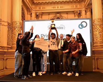 Estudiantes y docentes celebran en un escenario sosteniendo un trofeo de primer puesto y un diploma impreso frente a una pantalla