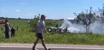 Restos de una avioneta ardiendo con humo blanco espeso en un campo de hierba verde junto a una carretera, bajo un cielo azul. Varias personas observan la escena