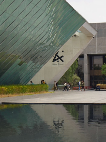Exterior del Museo Universitario Arte Contemporáneo con fachada inclinada de cristal verde, base de concreto y un estanque. Varias personas caminan frente al edificio