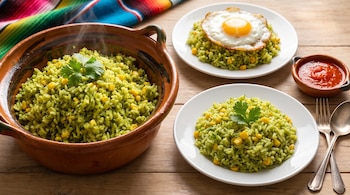 Cazuela de barro y platos con arroz verde y maíz, un plato con huevo frito, un cuenco de salsa roja y cubiertos sobre una mesa de madera.