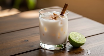 Primer plano de un vaso de agua de horchata con hielo y canela en polvo, con una rama de canela. Un medio limón verde y gotas de líquido están sobre una mesa de madera.