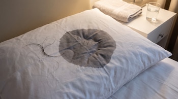 Almohada blanca con mancha circular oscura de humedad y hebras de cabello negro; en un velador, una toalla doblada y un vaso de agua.