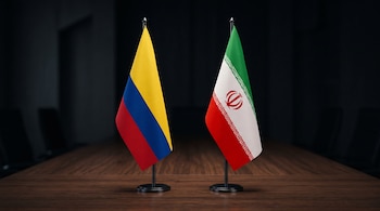 Dos pequeñas banderas de escritorio, una de Colombia y otra de Irán, ubicadas sobre una mesa de madera en una sala oscura.
