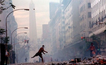Una calle con humo, escombros y fuego, personas, un hombre lanzando un objeto, edificios altos y el Obelisco de Buenos Aires al fondo