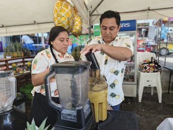 Hombre y mujer asiáticos sonriendo en primer plano, la mujer sostiene un letrero con forma de piña que dice "Feria de la Piña 2026 Santa María Ostuma"