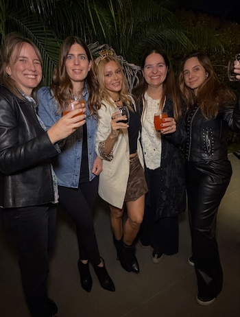 Cinco mujeres posan de pie en un espacio exterior durante la noche mientras sostienen copas con bebidas frente a plantas