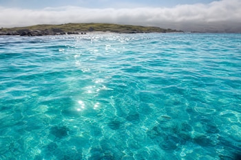 Agua de mar de color turquesa con reflejos de luz solar en primer plano y una colina verde bajo nubes al fondo.