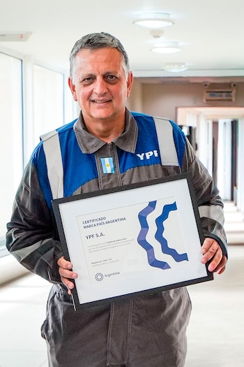 Hombre sonriente en uniforme de trabajo azul y gris, con un parche de la bandera argentina, sosteniendo un certificado enmarcado de YPF Marca País Argentina