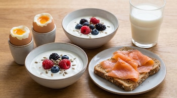 Vista aérea de un desayuno con dos huevos pasados por agua, dos cuencos de yogur con frutos rojos y semillas, tostadas con salmón ahumado y un vaso de leche.