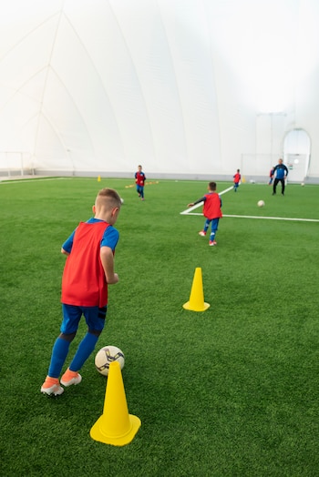 Niño con camiseta roja y uniforme azul juega fútbol con una pelota cerca de un cono amarillo. Otros niños y un adulto en cancha cubierta de pasto sintético