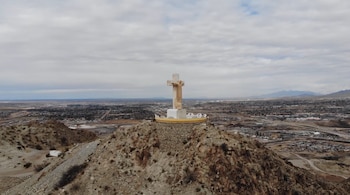 Estatua de Cristo Rey de color crema sobre una colina rocosa, con una ciudad extensa y montañas lejanas bajo un cielo cubierto de nubes