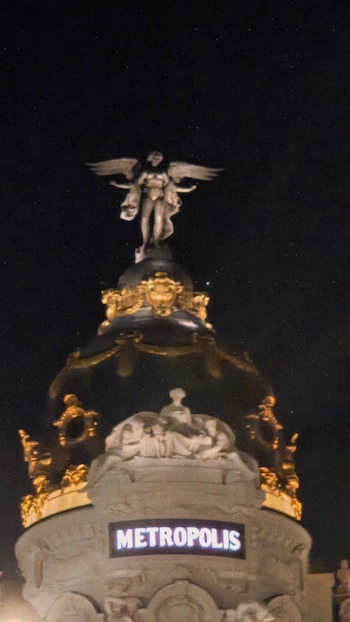 Cúpula oscura con detalles dorados, una estatua alada en la cima y un cartel con la palabra METROPOLIS iluminada de noche