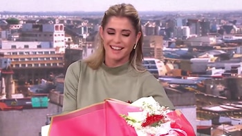 Pamela David sonríe con una expresión de sorpresa y alegría mientras sostiene un ramo de flores rojas y blancas, con un fondo urbano de edificios