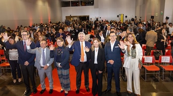 Un grupo de personas en traje saluda con la mano al frente de un auditorio lleno de asistentes que lanzan birretes al aire