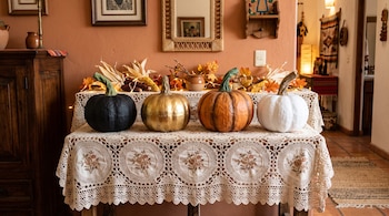 Cuatro calabazas decorativas de color negro, dorado, naranja y blanco sobre un mantel de encaje blanco con luces y elementos de otoño al fondo.