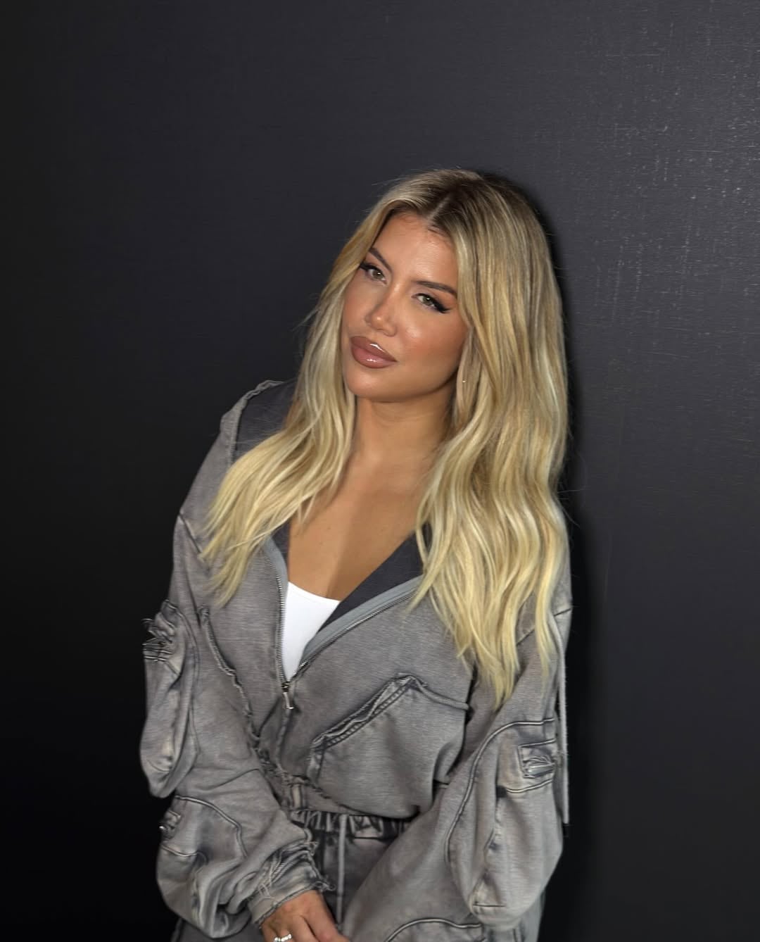 Wanda Nara posa con un conjunto deportivo gris deslavado, cabello rubio suelto y un maquillaje sofisticado que resalta sus labios y ojos.