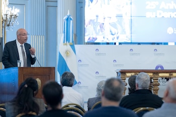 Hombre con traje y gafas habla en un podio con micrófono, junto a la bandera argentina. El público asiste de espaldas y una pantalla con logotipos de Legislatura es visible
