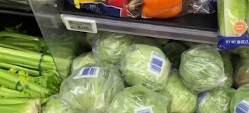 Cabezas de lechuga empaquetadas en bolsas de plástico, manojos de apio atados, pimientos envasados y una etiqueta de precio de 6.00 en un estante de supermercado