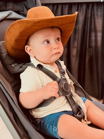 Un bebé sentado en una silla de paseo con un sombrero cowboy marrón, camisa clara y shorts de mezclilla, con un arnés de seguridad