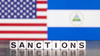 Vista de cerca de cubos de letras que deletrean "SANCTIONS" sobre una superficie reflectante, con las banderas de Estados Unidos y Nicaragua desenfocadas al fondo.