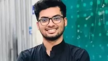 Um jovem de barba, óculos e camisa preta sorri diretamente para a câmera. O fundo está desfocado com tons brancos e verdes