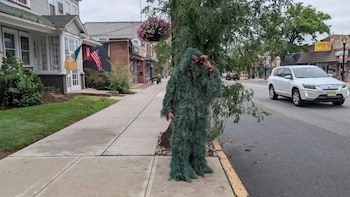 Persona con traje ghillie verde oscuro sostiene binoculares en una acera, junto a un árbol; vehículos y edificios se observan al fondo